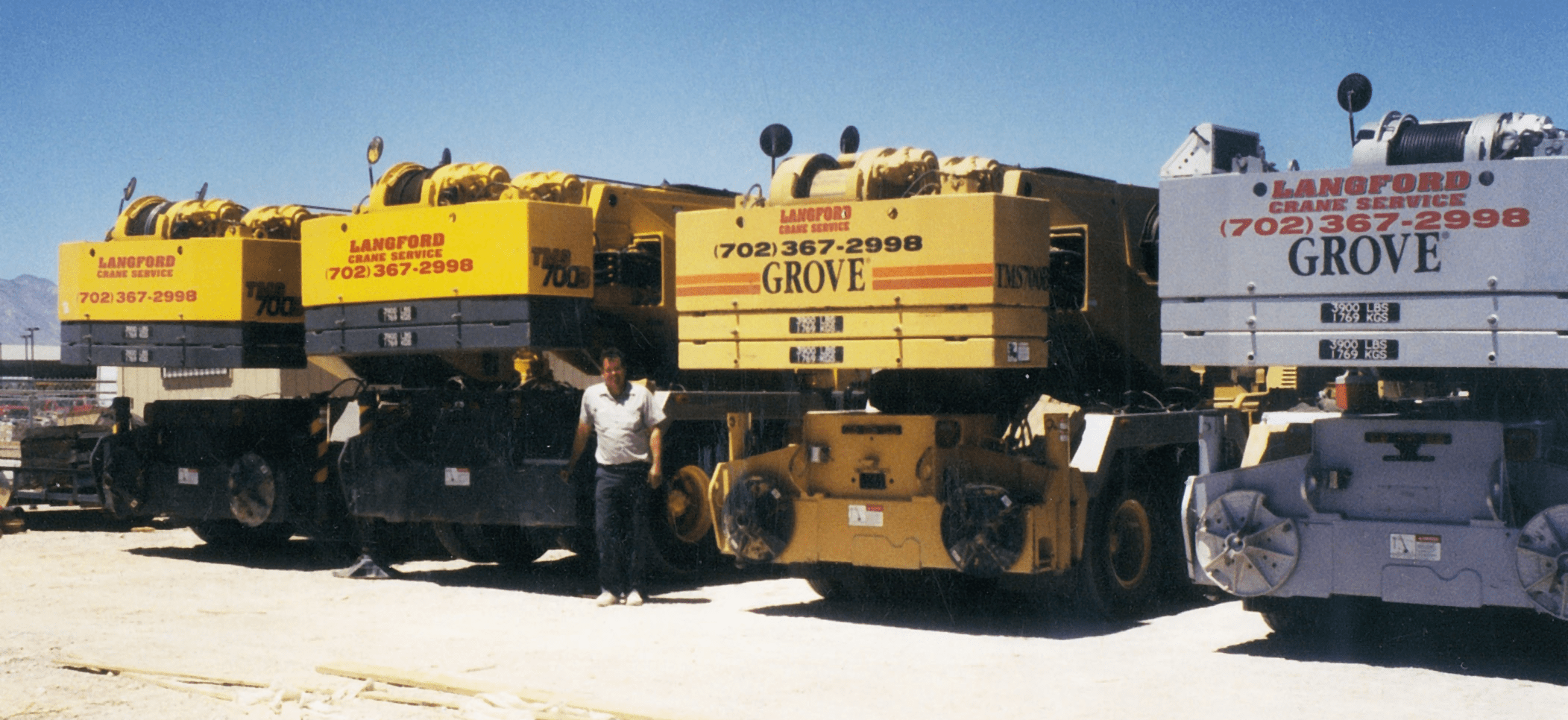 Four large cranes in a construction yard, labeled "Langford Crane Service" and "Grove." A man stands between the cranes, showcasing equipment used for heavy lifting.