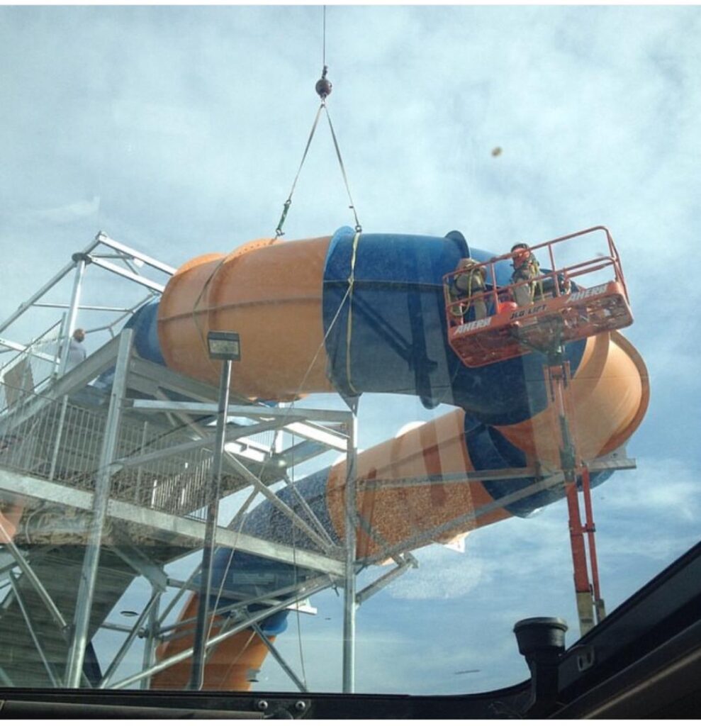 Workers on a lift are repairing a large orange and blue water slide at an amusement park under a partly cloudy sky.