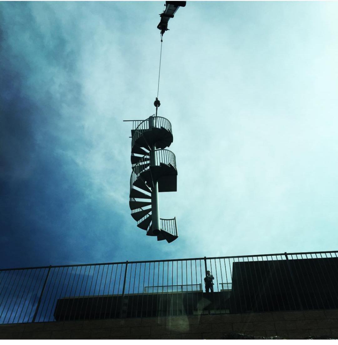 A spiral staircase suspended from a crane, silhouetted against a cloudy sky, with a person standing below observing the installation process.
