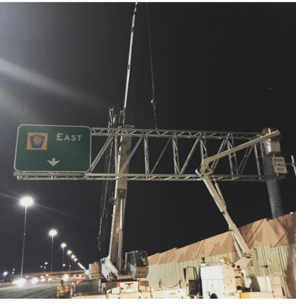 Construction crew installing an overhead highway sign indicating "East" during nighttime. Safety equipment and cranes are visible in the scene.
